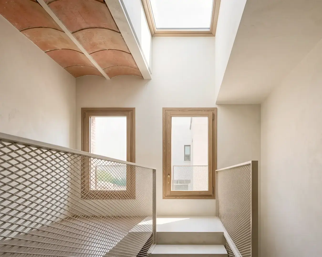 Núcleo de escalera de vivienda reformada en Badalona, con lucernario cenital, techo de bóveda catalana de rasilla vista, barandilla de malla metálica y carpinterías de madera. Proyecto de Tova Arquitectura