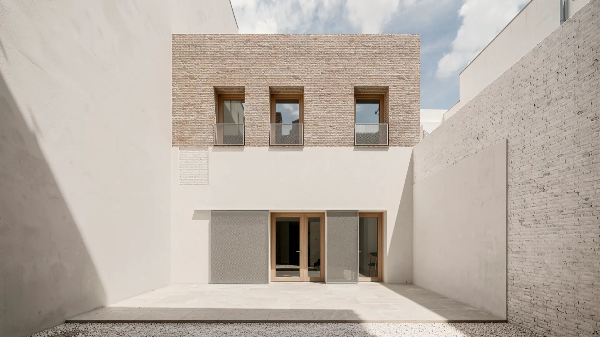 Patio interior de vivienda reformada en Badalona, con pavimento de grava, fachada encalada, carpinterías de madera y ampliación superior en ladrillo cara vista. Proyecto de Tova Arquitectura