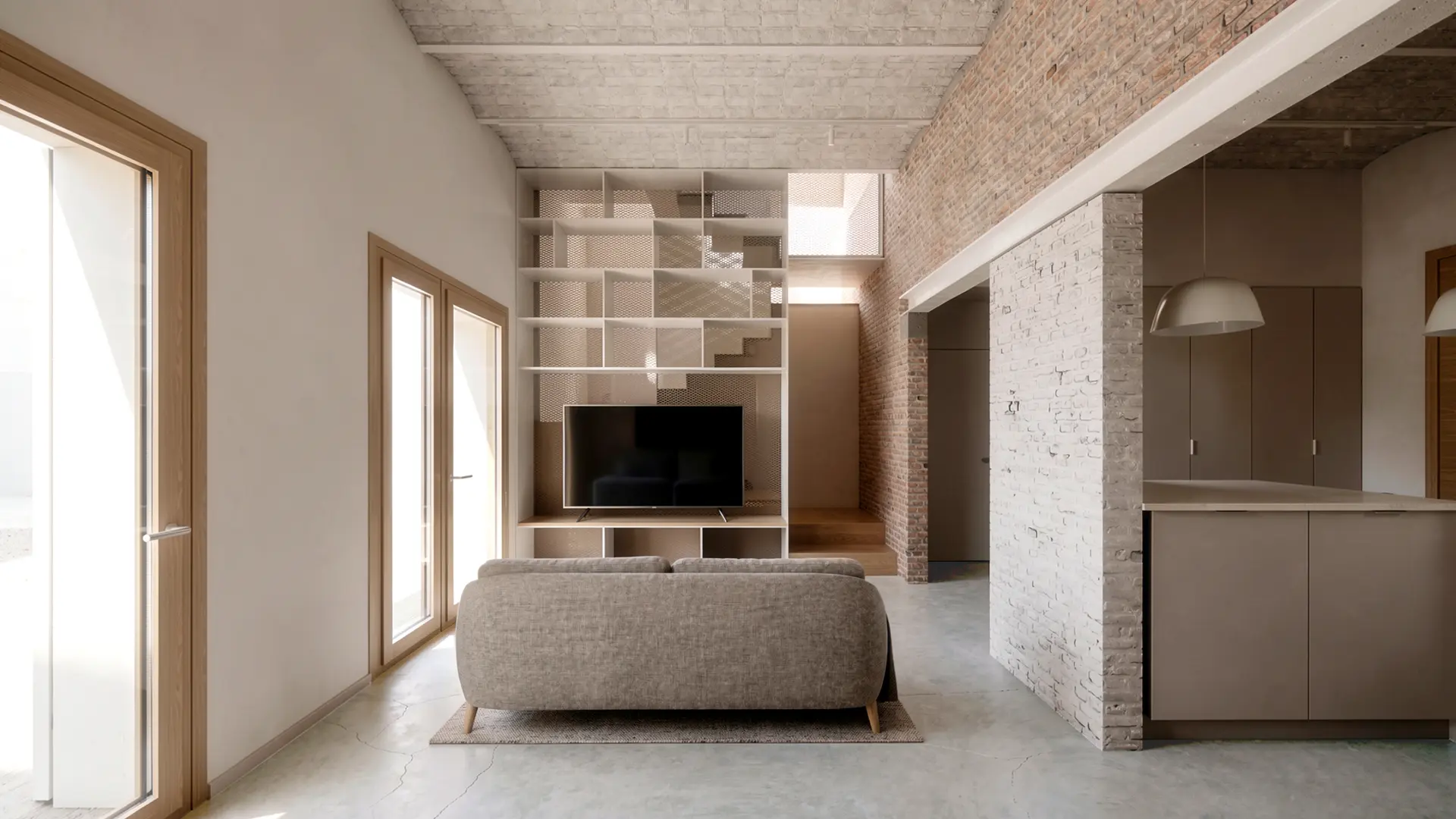 Salón de vivienda reformada en Badalona, con librería a doble altura integrada en el núcleo de escalera, ladrillo antiguo recuperado visto, suelo de microcemento y carpinterías de madera. Proyecto de Tova Arquitectura