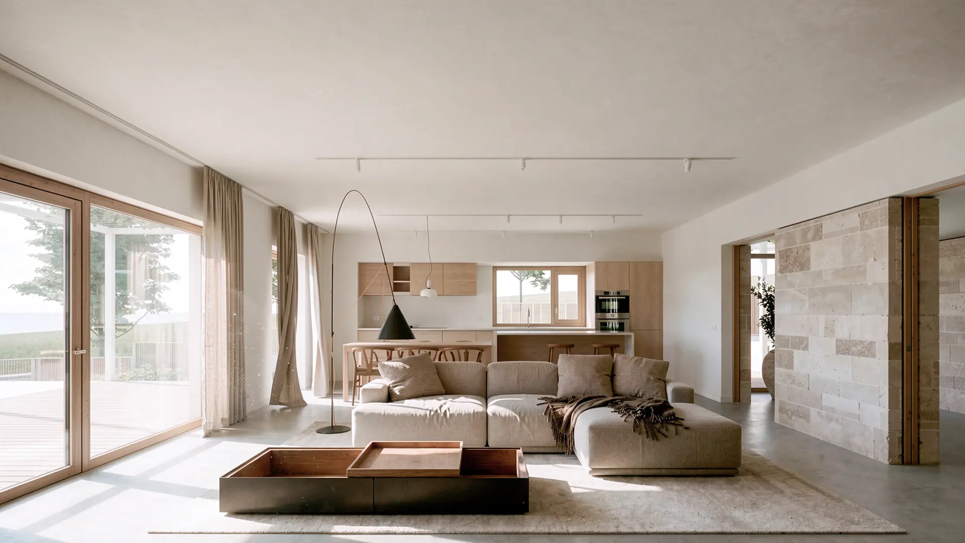 Salón comedor en planta abierta de vivienda unifamiliar de obra nueva en L'Ametlla del Vallès, con suelo de microcemento, muro interior de piedra natural, cocina integrada de madera y grandes ventanales con vistas al paisaje. Proyecto de Tova Arquitectura