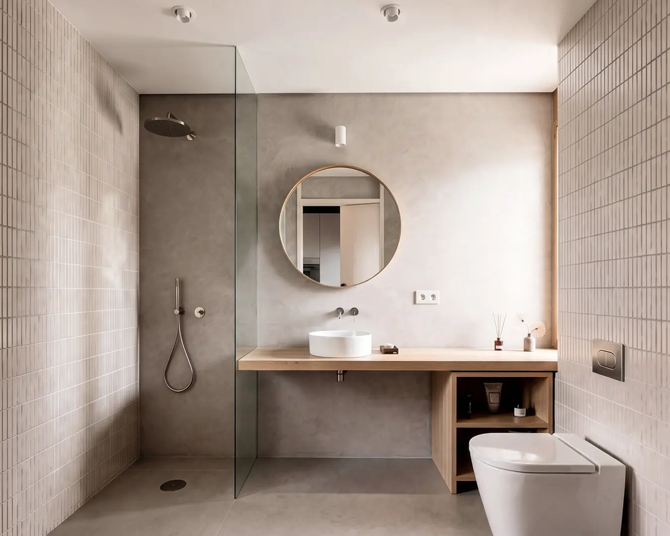 Baño de vivienda reformada en Badalona, con ducha italiana de microcemento, mampara de vidrio, azulejo rectangular en tono crema, mueble de madera con lavabo sobre encimera y espejo redondo. Proyecto de Tova Arquitectura.