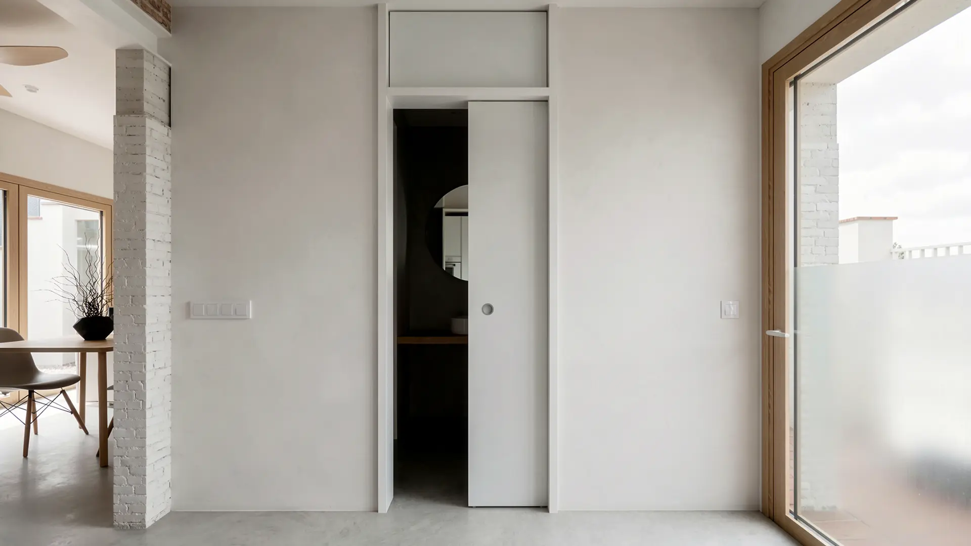 Acceso al baño de vivienda reformada en Badalona, con puerta corredera lacada en blanco integrada en el paramento, ladrillo antiguo recuperado visto y suelo de microcemento. Proyecto de Tova Arquitectura