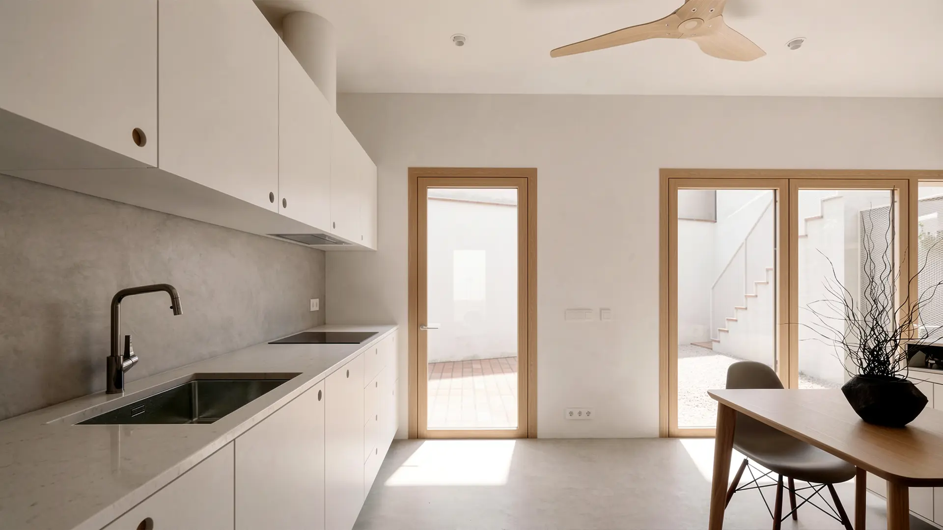 Cocina de vivienda reformada en Badalona, con muebles lacados en blanco, encimera y salpicadero de microcemento, carpinterías de madera y apertura directa al patio interior. Proyecto de Tova Arquitectura