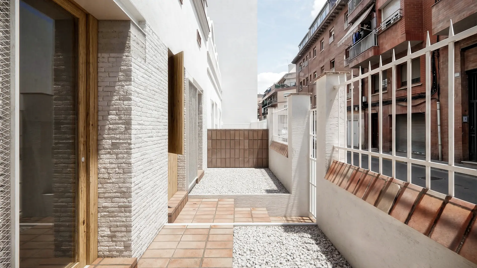 Patio de acceso de vivienda reformada en Badalona, con pavimento de barro, grava blanca, ladrillo antiguo recuperado visto y carpinterías de madera en entorno urbano consolidado. Proyecto de Tova Arquitectura
