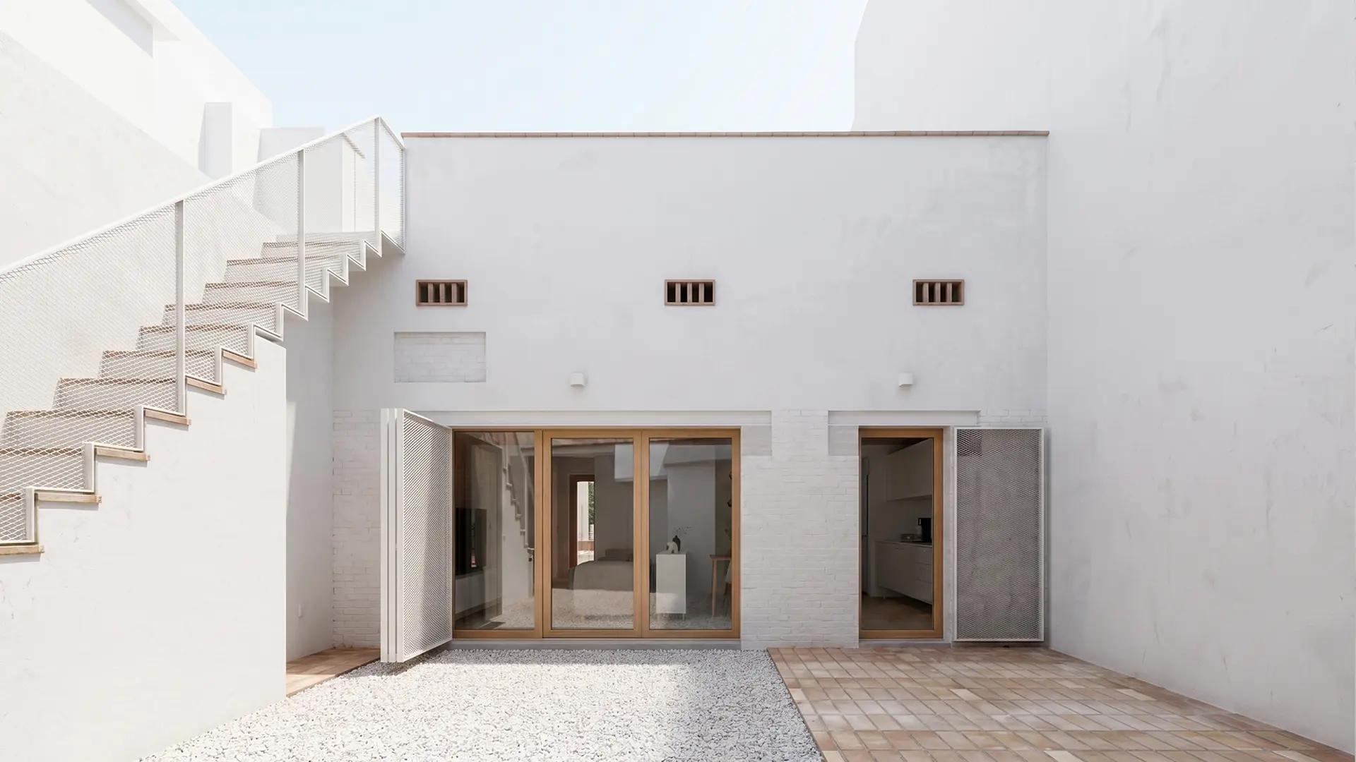 Patio interior de vivienda reformada en Badalona, con fachada encalada, pavimento de barro y grava blanca, carpinterías de madera y escalera exterior con barandilla de malla metálica. Proyecto de Tova Arquitectura