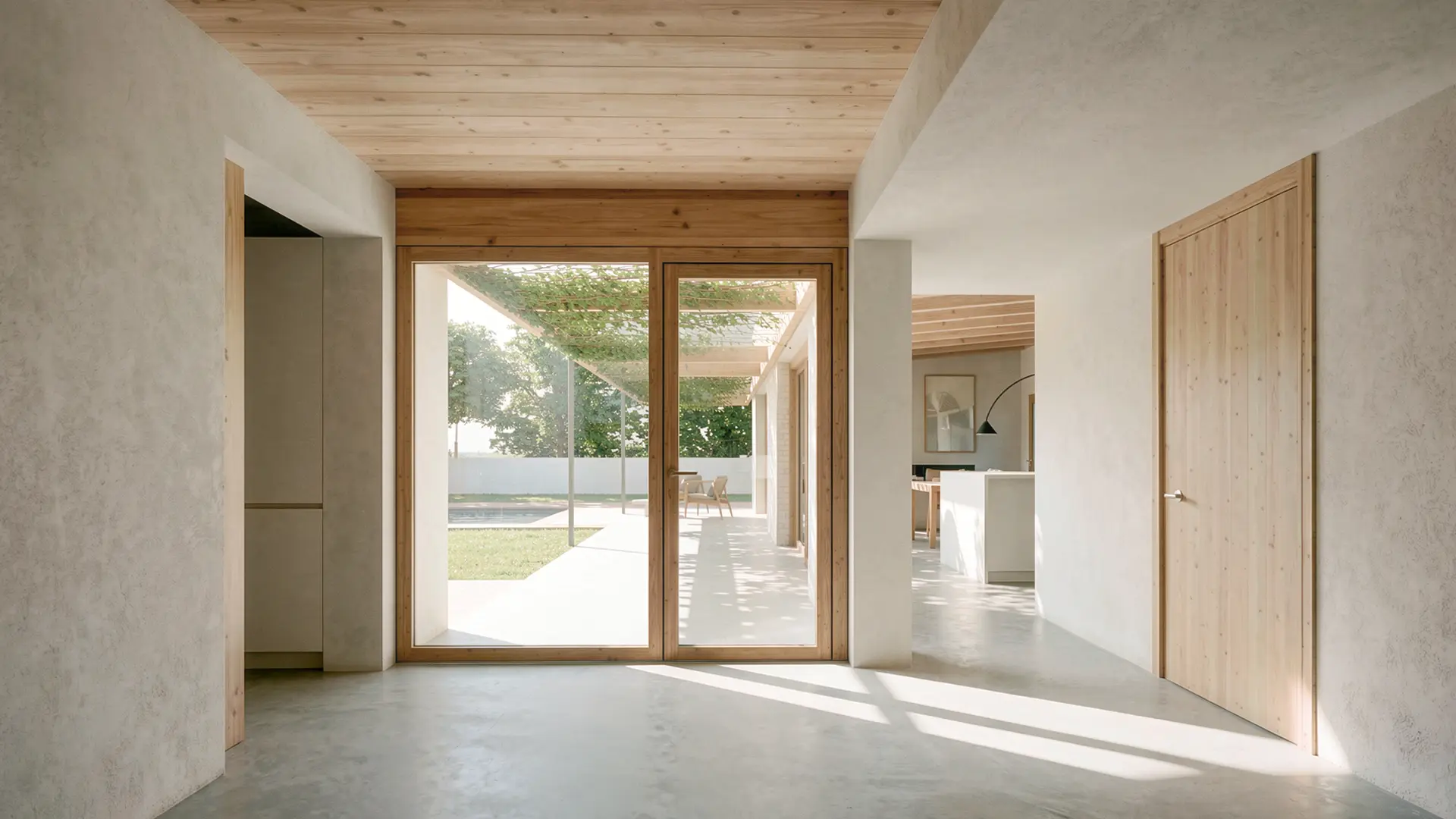 Entrada interior de vivienda unifamiliar de obra nueva en Cardedeu, con suelo de microcemento, techo de madera vista y puerta de madera que conecta visualmente el interior con el jardín y la piscina. Proyecto de Tova Arquitectura