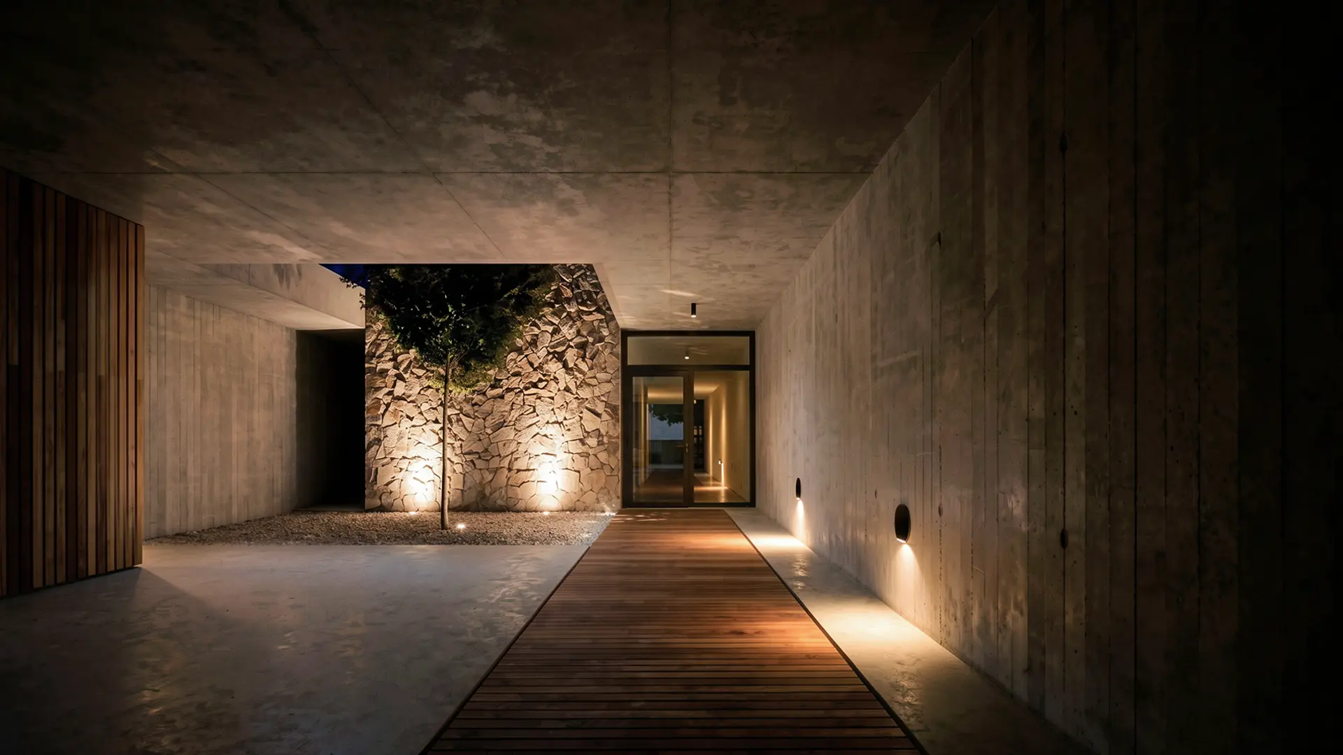 Acceso de noche a vivienda unifamiliar de obra nueva en Cardedeu, con pasarela de tarima de madera iluminada entre paredes de hormigón visto y patio interior con árbol y muro de mampostería de piedra natural. Propuesta de diseño de Tova Arquitectura