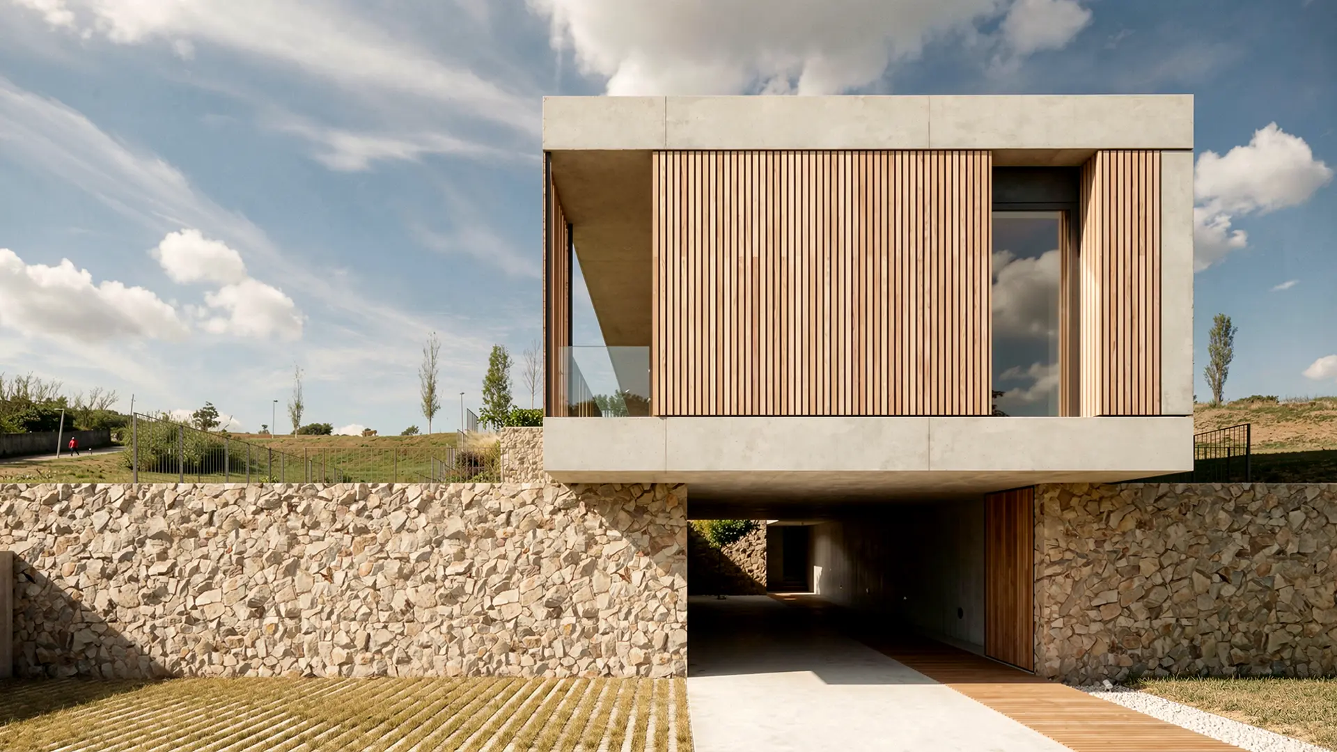 Fachada principal de vivienda unifamiliar de obra nueva en Cardedeu, con volumen superior de hormigón visto y lamas de madera sobre zócalo de mampostería de piedra natural. Propuesta de diseño de Tova Arquitectura.