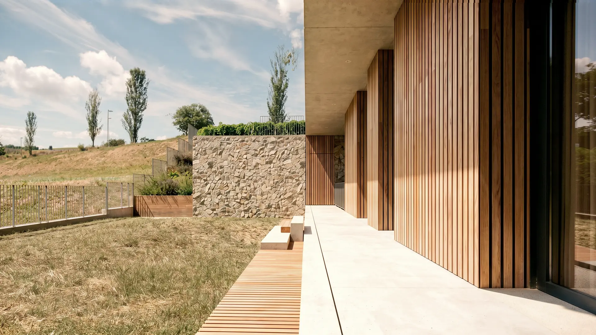 Terraza y jardín de vivienda unifamiliar de obra nueva en Cardedeu, con tarima de madera, lamas de madera verticales en fachada, muro de mampostería de piedra natural y vistas al paisaje del Vallès Oriental. Propuesta de diseño de Tova Arquitectura